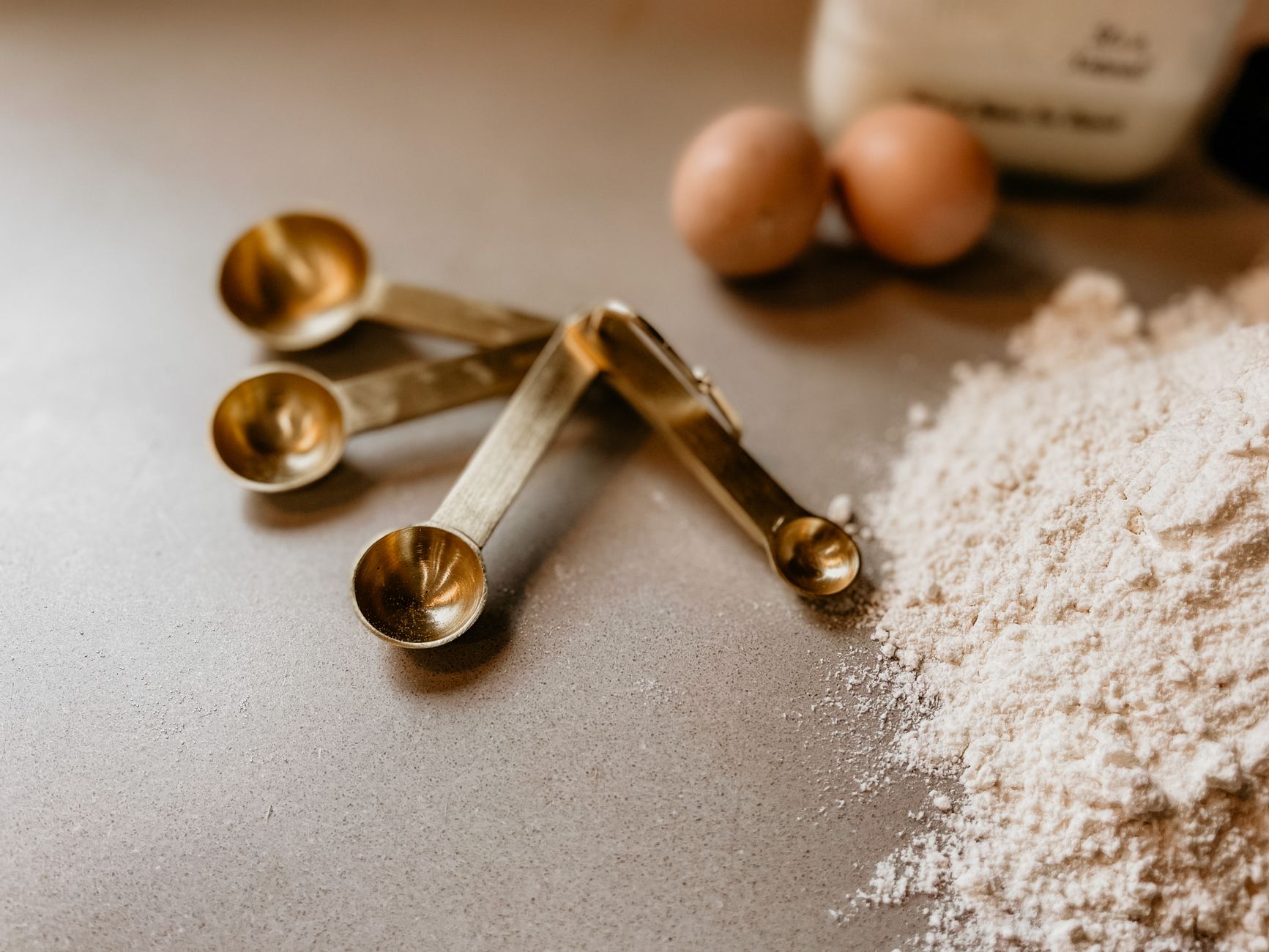 Trzy złote łyżki miarowe obok mąki i jaj na kuchennym blacie