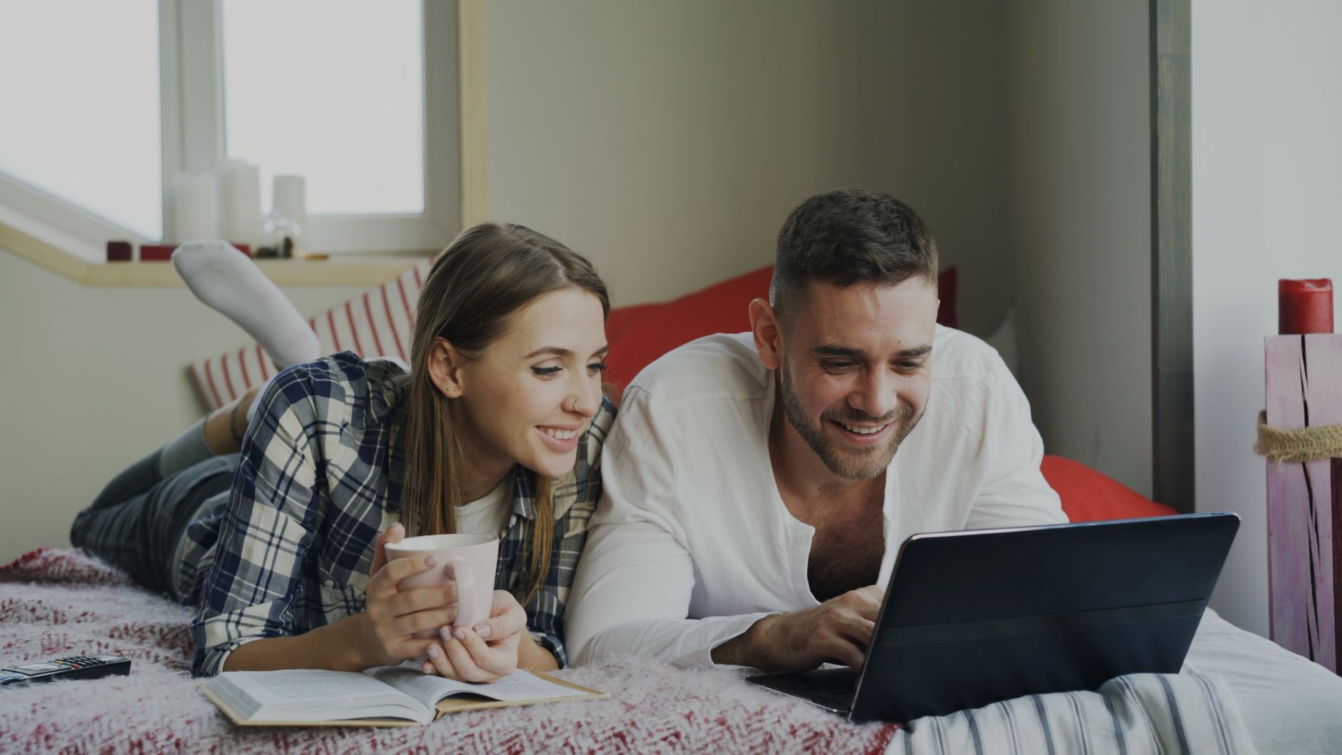 Para leżąca na łóżku patrzy razem na tablet, kobieta trzyma kubek, oboje się uśmiechają