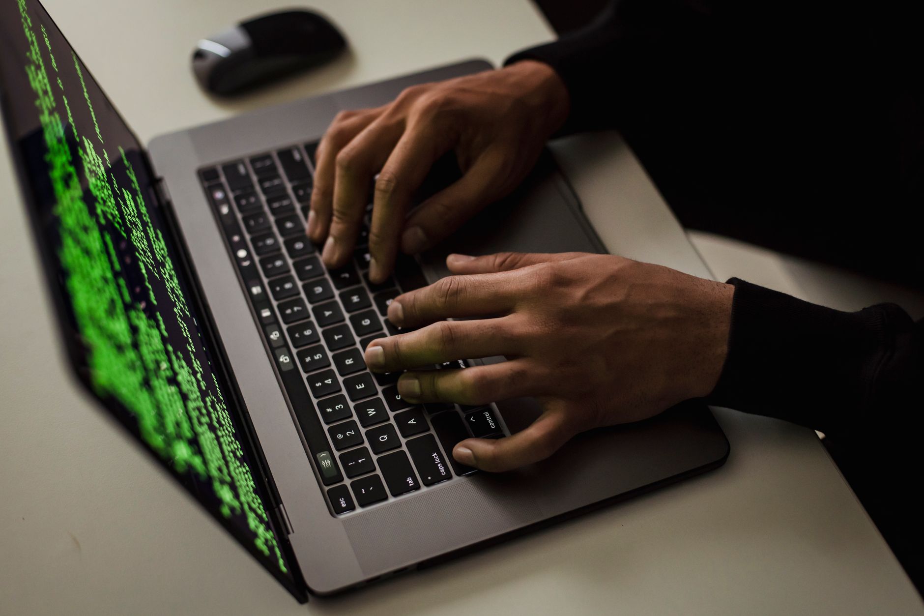 Ręce na klawiaturze laptopa z zielonym kodem na ekranie, na białym biurku z myszką w tle